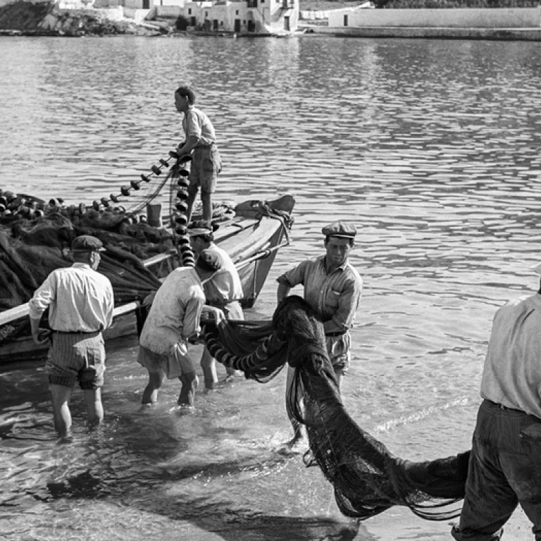 Μύκονος, 1955. Ξημερώματα στον μόλο Καμπάνη, ψαράδες ξεφορτώνουν τα δίχτυα τους για στέγνωμα και επισκευή. Το νεόκτιστο ξενοδοχείο Λητώ διαφαίνεται στο βάθοςi